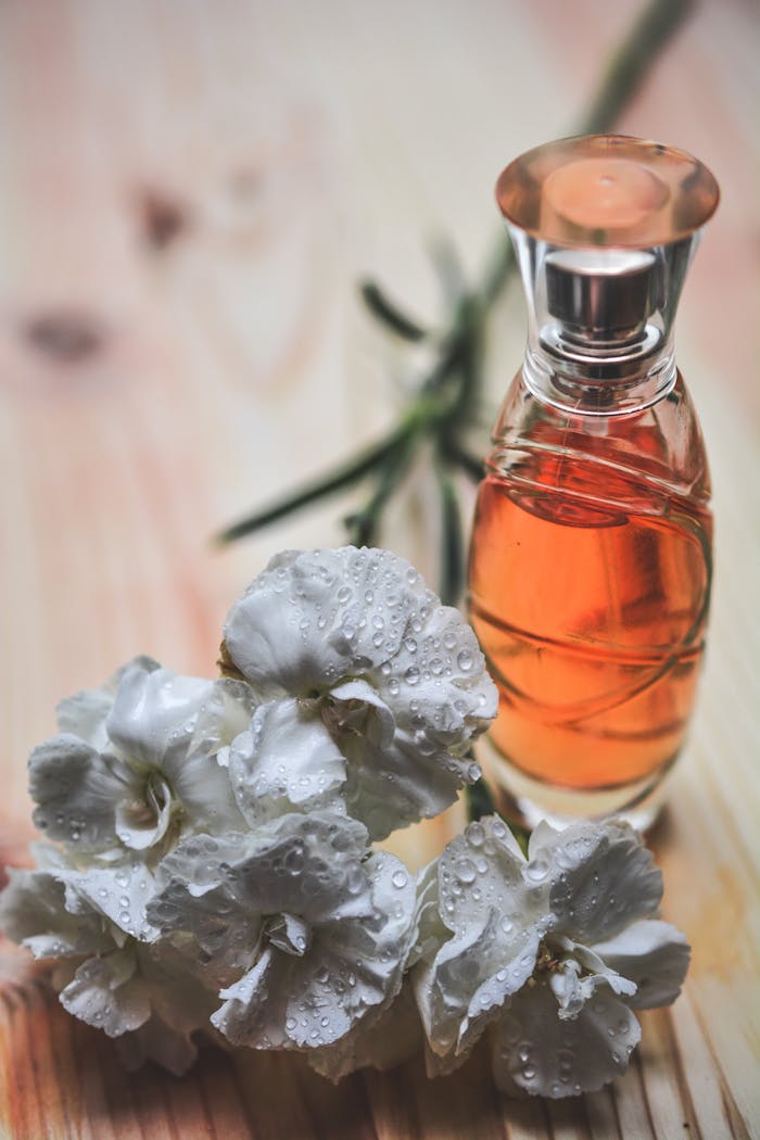 our-story Glass perfume bottle alongside dew-kissed white carnation flowers on wood surface.